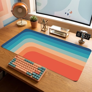 Puede incluir: Un escritorio con una alfombrilla de ratón de estilo retro con un diseño de arcoíris en tonos azules, naranjas y rojos. Un teclado color melocotón con teclas turquesas está sobre la alfombrilla. El escritorio también incluye una lámpara de escritorio, una planta suculenta y un reloj digital.