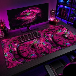 May include: A large, rectangular mouse mat featuring a striking pink snakeskin design. The pattern has a detailed, textured look with green and blue accents. A black keyboard and mouse sit on the mat, with a computer monitor visible in the background.