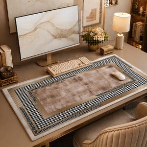 Puede incluir: Alfombrilla de escritorio con un centro marrón y un borde de pata de gallo en blanco y negro. La alfombrilla tiene un borde dorado y está rodeada por un monitor de ordenador, un teclado y un ratón. El escritorio es de color marrón claro.