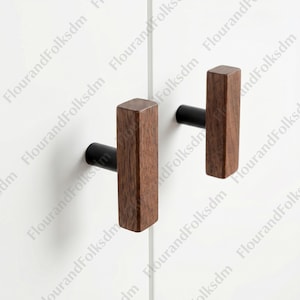 May include: Two wooden cabinet pulls with a dark brown finish and a black cylindrical base. The pulls are rectangular and have a modern, minimalist design. The pulls are mounted on a white cabinet door.