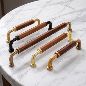 May include: A collection of cabinet pulls with wooden handles and metal accents. The pulls feature a dark wood grain center with curved metal ends in gold and black finishes. The hardware is arranged on a white marble surface.