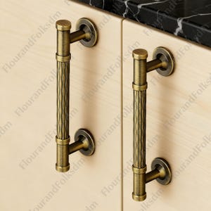 May include: Two antique brass cabinet pulls with a vertical, textured design. The pulls are mounted on a light-coloured wooden cabinet door. The hardware has a classic, vintage aesthetic, suitable for kitchen or bathroom cabinetry.
