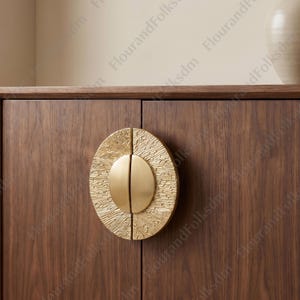 May include: A close-up of a wooden cabinet door with a gold-colored, textured oval handle. The handle is split into two halves with a smooth, rounded center. The cabinet door is a dark brown color.