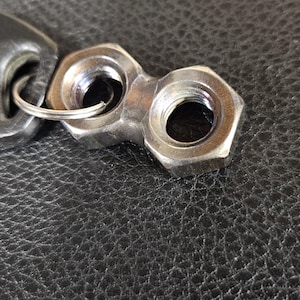 May include: Two silver-colored hexagonal metal nuts joined together to form a single piece. A small, circular silver metal split ring is threaded through the opening of one of the hexagonal nuts, connecting it to another unseen metal object.