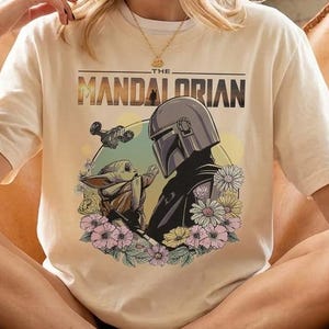 以下が含まれることがあります： 「THE MANDALORIAN」の文字と、マンダロリアンとグローグーが花に囲まれたイラストが描かれたクリーム色のTシャツ。茶色の革張りの椅子に座っている人が着ています。
