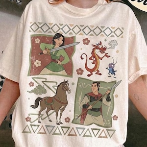 Może przedstawiać: Koszulka w kolorze kości słoniowej z nadrukiem graficznym przedstawiającym postacie z filmu Mulan, w tym wojowniczkę z mieczem, czerwonego smoka, małego niebieskiego owada, brązowego konia i wojownika z włócznią, otoczonych geometrycznymi wzorami.