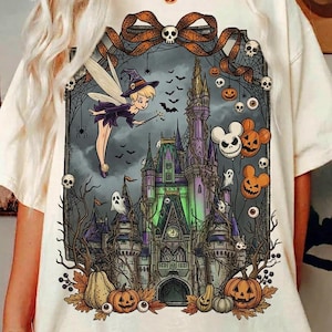 May include: A cream-colored t-shirt featuring a graphic print of a fairy in a witch hat flying toward a castle. The design includes spiderwebs, skulls, pumpkins, and mouse-shaped jack-o'-lanterns, all framed by a large orange bow at the top.