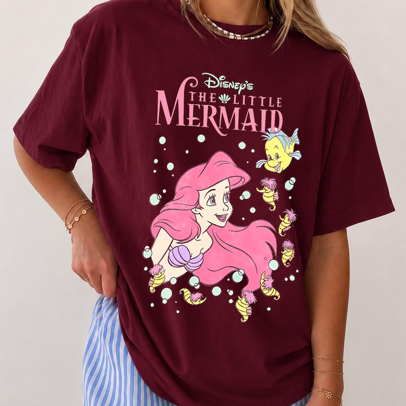 May include: A maroon short-sleeved t-shirt featuring a graphic print of Ariel from Disney's The Little Mermaid with pink hair, purple seashell top, and yellow sea creatures. The text reads Disney's The Little Mermaid in white lettering.