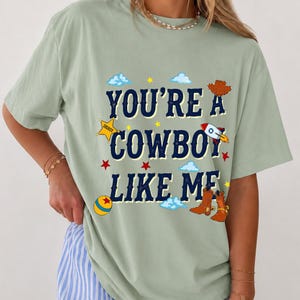 Puede incluir: Una camiseta de manga corta color verde salvia con el texto en azul oscuro "YOU'RE A COWBOY LIKE ME" rodeado de gr&aacute;ficos coloridos que incluyen una estrella de sheriff amarilla, un cohete, una pelota redonda, botas vaqueras marrones, estrellas rojas y nubes azules.