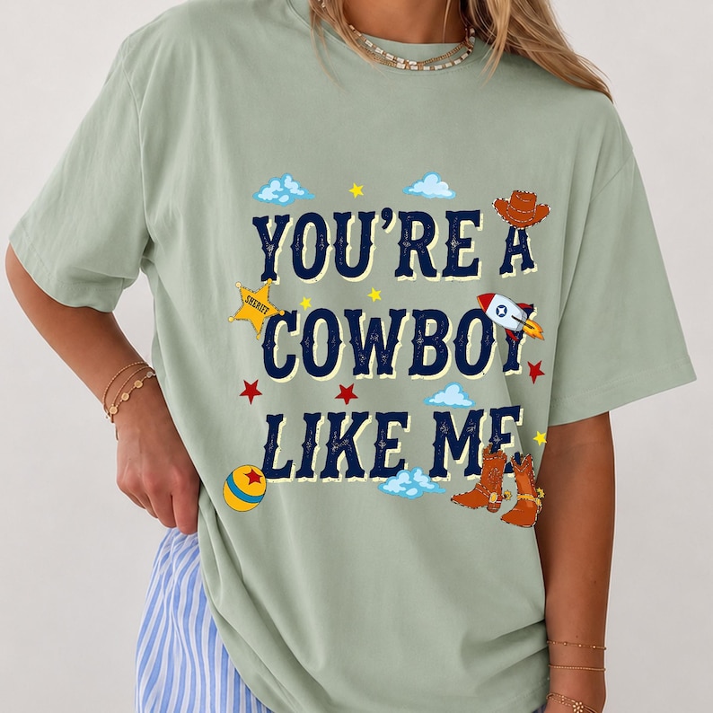 Puede incluir: Una camiseta de manga corta color verde salvia con el texto en azul oscuro "YOU'RE A COWBOY LIKE ME" rodeado de gr&aacute;ficos coloridos que incluyen una estrella de sheriff amarilla, un cohete, una pelota redonda, botas vaqueras marrones, estrellas rojas y nubes azules.
