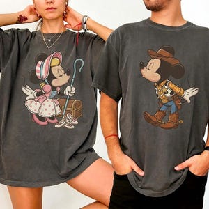 May include: Two charcoal gray short-sleeve t-shirts featuring graphic prints. The left shirt displays a character dressed as Bo Peep with a bonnet and staff. The right shirt shows a character dressed as a cowboy with a hat, vest, and boots.