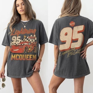 K&ouml;nnte beinhalten: Dunkelgraues T-Shirt mit einer Grafik von Lightning McQueen aus dem Film Cars. Auf der Vorderseite steht "Lightning 95 McQueen" mit einem roten Rennwagen. Auf der R&uuml;ckseite steht gro&szlig; "95" und der Text "Lightning McQueen".
