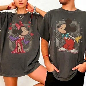 May include: Two charcoal grey t-shirts featuring graphic prints. The left shirt displays Minnie Mouse in a red sequined dress with purple gloves and red hearts. The right shirt shows Mickey Mouse in red trousers and a blue polka-dot bow tie.
