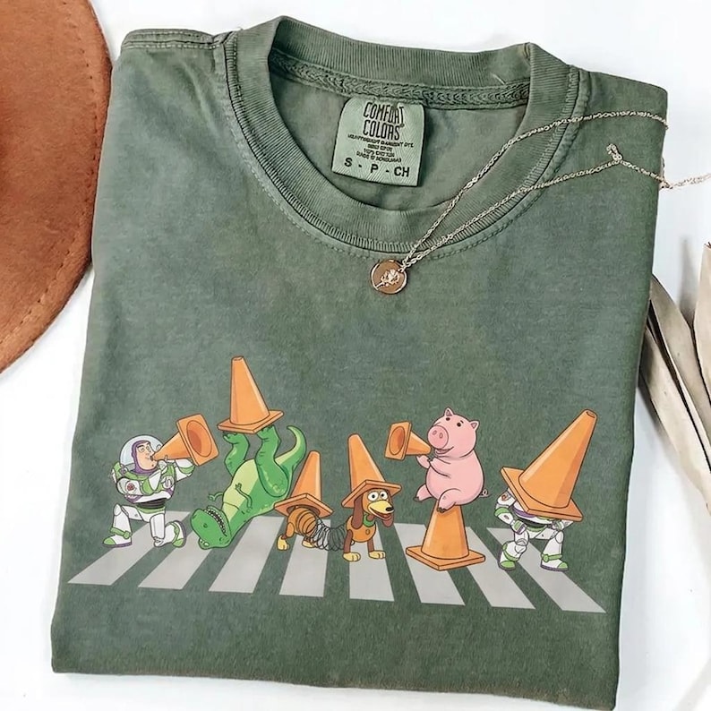 May include: Green Comfort Colors t-shirt featuring a graphic of cartoon characters wearing orange traffic cones and crossing a crosswalk. The characters include Buzz Lightyear, a dinosaur, a dachshund, and a pig. A silver necklace with a pendant is also visible.