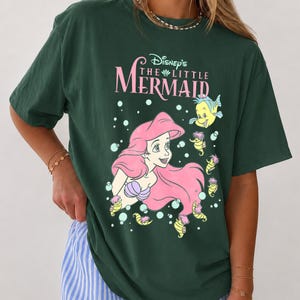 May include: A dark green short-sleeve t-shirt featuring a graphic print of Ariel from Disney's The Little Mermaid with pink hair, purple shells, and yellow sea creatures. The text reads Disney's The Little Mermaid in pink and white lettering.