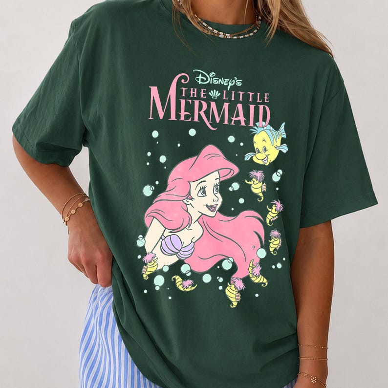 May include: A dark green short-sleeve t-shirt featuring a graphic print of Ariel from Disney's The Little Mermaid with pink hair, purple shells, and yellow sea creatures. The text reads Disney's The Little Mermaid in pink and white lettering.