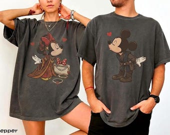 May include: Two charcoal gray short-sleeve t-shirts featuring graphic prints of cartoon characters. The left shirt displays a character in a red dress with a bow, while the right shirt shows a character in a dark tunic, both with small red hearts.