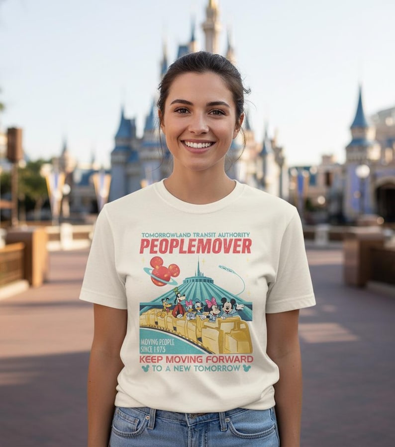 May include: A cream t-shirt with a graphic of the Tomorrowland Transit Authority PeopleMover ride. The design includes Mickey Mouse and friends riding the PeopleMover, with text that reads "PEOPLEMOVER" and "KEEP MOVING FORWARD."