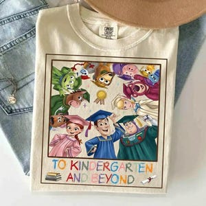 Puede incluir: Camiseta color crema con un gr&aacute;fico colorido de personajes de Toy Story, incluyendo a Woody y Buzz Lightyear, con atuendo de graduaci&oacute;n. El texto "TO KINDERGARTEN AND BEYOND" se muestra debajo. La camiseta es de material suave.