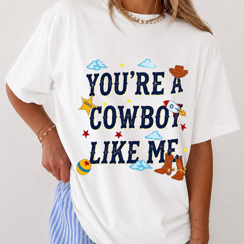 Puede incluir: Una camiseta blanca con el texto azul oscuro "YOU'RE A COWBOY LIKE ME" rodeado de gr&aacute;ficos coloridos que incluyen una estrella de sheriff amarilla, un sombrero de vaquero marr&oacute;n, un cohete, una pelota amarilla y azul, botas y nubes.