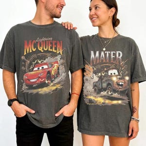 May include: Two charcoal grey t-shirts featuring characters from the movie Cars. One shirt displays Lightning McQueen with the text "Lightning McQueen". The other shirt features Mater with the text "Mater". Both shirts have a vintage, distressed look.