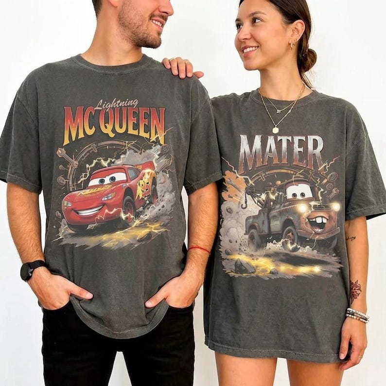 May include: Two charcoal grey t-shirts featuring characters from the movie Cars. One shirt displays Lightning McQueen with the text "Lightning McQueen". The other shirt features Mater with the text "Mater". Both shirts have a vintage, distressed look.