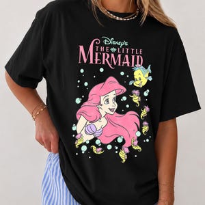 May include: A black short-sleeved t-shirt featuring a graphic print of Ariel from Disney's The Little Mermaid with pink hair, a purple seashell top, and Flounder the fish. The text reads Disney's The Little Mermaid in pink and white lettering.