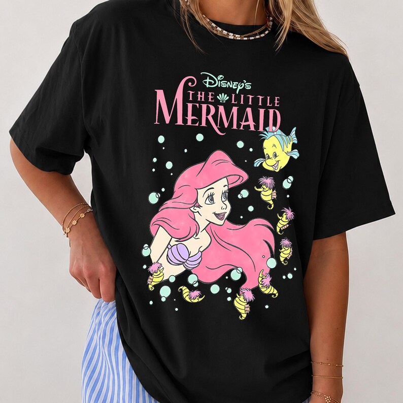 May include: A black short-sleeved t-shirt featuring a graphic print of Ariel from Disney's The Little Mermaid with pink hair, a purple seashell top, and Flounder the fish. The text reads Disney's The Little Mermaid in pink and white lettering.