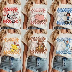Pode incluir: Seis camisetas creme com gr&aacute;ficos de desenhos animados. Cada camisa apresenta um nome e imagem de personagem diferente, incluindo Duke Caboom, Hamm, Giggle McDimples, Ducky, Gabby e Ken, com um fundo quadriculado e o ano de 1995.
