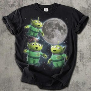 Może przedstawiać: Czarny t-shirt z krótkim rękawem z nadrukiem graficznym przedstawiającym trzech zielonych, trzyokich kosmitów w skafandrach kosmicznych unoszących się przed dużym, szczegółowym księżycem. Tło to ciemne, rozgwieżdżone niebo z widocznymi gwiazdami.