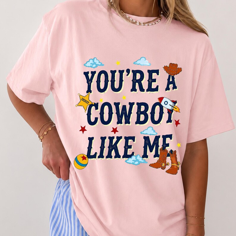 Puede incluir: Una camiseta rosa claro con el texto azul oscuro "YOU'RE A COWBOY LIKE ME" rodeado de ilustraciones gr&aacute;ficas de una estrella de sheriff amarilla, un sombrero de vaquero marr&oacute;n, un cohete, una pelota colorida, estrellas rojas y botas.