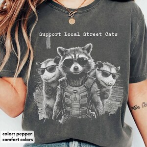 Peut inclure: Un t-shirt gris anthracite avec un motif représentant un raton laveur portant un gilet tactique et deux opossums, tous portant des lunettes de soleil noires. Le texte Support Local Street Cats est imprimé en blanc au-dessus des animaux.