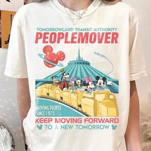 May include: Cream-coloured t-shirt with a vibrant graphic of the Tomorrowland Transit Authority PeopleMover ride. The design features Mickey Mouse, Minnie Mouse, Goofy, Donald Duck, and Daisy Duck. Text includes "PEOPLEMOVER" and "MOVING PEOPLE SINCE 1975".