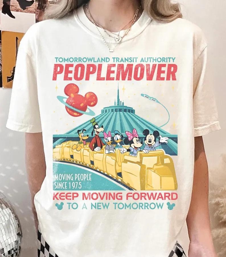 May include: Cream-coloured t-shirt with a vibrant graphic of the Tomorrowland Transit Authority PeopleMover ride. The design features Mickey Mouse, Minnie Mouse, Goofy, Donald Duck, and Daisy Duck. Text includes "PEOPLEMOVER" and "MOVING PEOPLE SINCE 1975".