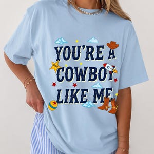 Puede incluir: Una camiseta azul claro con el texto azul oscuro "YOU'RE A COWBOY LIKE ME" rodeado de ilustraciones gr&aacute;ficas de una estrella de sheriff amarilla, un cohete, un sombrero de vaquero marr&oacute;n, botas de vaquero marrones, una pelota y nubes.