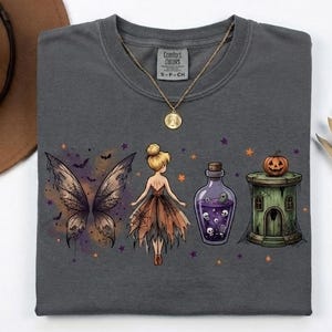 Puede incluir: Una camiseta gris carbón con un estampado gráfico de alas de hada, una figura de hada, una botella de poción morada llena de calaveras y una torre verde coronada con una calabaza. Un collar con colgante dorado descansa sobre el cuello.