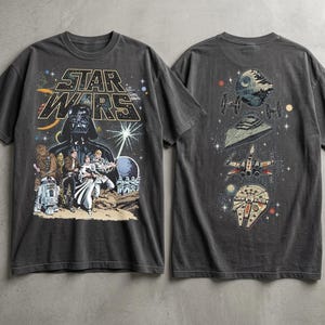Pode incluir: T-shirt cinza escuro com um gráfico de Star Wars na frente, com Darth Vader e outros personagens. A parte de trás da camisa mostra vários veículos espaciais. O design inclui o logotipo de Star Wars e elementos espaciais.