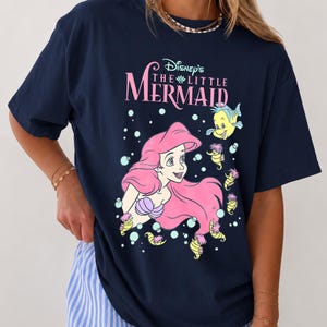 May include: A navy blue t-shirt featuring a graphic print of Ariel from Disney's The Little Mermaid with pink hair, a purple seashell top, and yellow sea creatures. The text reads Disney's The Little Mermaid in pink and white lettering.