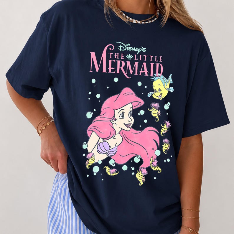 May include: A navy blue t-shirt featuring a graphic print of Ariel from Disney's The Little Mermaid with pink hair, a purple seashell top, and yellow sea creatures. The text reads Disney's The Little Mermaid in pink and white lettering.