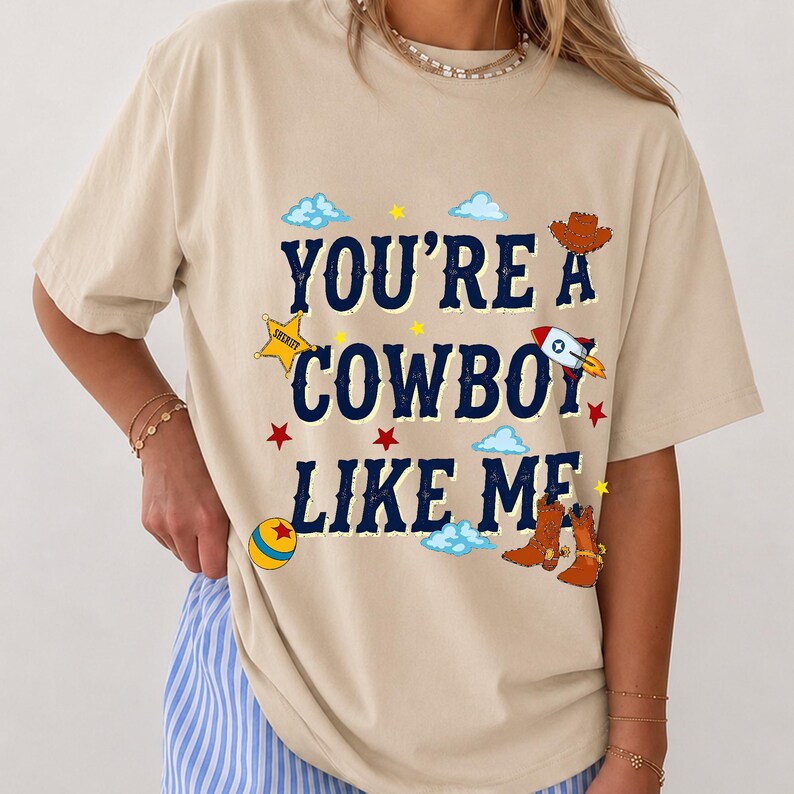 Puede incluir: Una camiseta beige de manga corta con el texto azul oscuro "YOU'RE A COWBOY LIKE ME" rodeado de gr&aacute;ficos coloridos que incluyen una estrella de sheriff amarilla, un cohete, un sombrero de vaquero marr&oacute;n, botas marrones, una pelota y nubes azules.