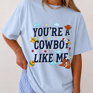 Puede incluir: Una camiseta azul claro con el texto azul oscuro "YOU'RE A COWBOY LIKE ME" rodeado de ilustraciones gr&aacute;ficas de un sombrero de vaquero marr&oacute;n, una estrella de sheriff amarilla, un cohete, una pelota colorida, estrellas rojas y nubes azul claro.