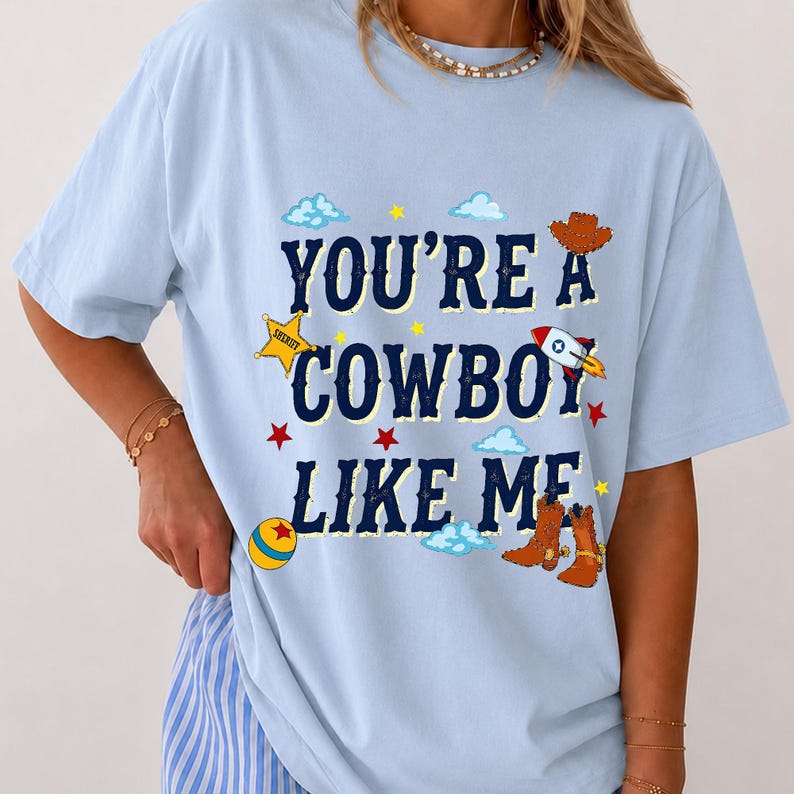 Puede incluir: Una camiseta azul claro con el texto azul oscuro "YOU'RE A COWBOY LIKE ME" rodeado de ilustraciones gr&aacute;ficas de un sombrero de vaquero marr&oacute;n, una estrella de sheriff amarilla, un cohete, una pelota colorida, estrellas rojas y nubes azul claro.