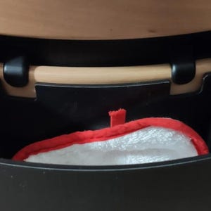 Puede incluir: Un contenedor de almacenamiento negro con un asa de madera y un guante de cocina rojo y blanco en el interior. El contenedor est&aacute; montado debajo de una superficie de madera y el guante tiene un borde rojo.