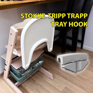 Op de afbeelding: Een Stokke Tripp Trapp kinderstoel met een wit dienblad en een grijze dienbladhaak. De kinderstoel is gemaakt van licht hout en heeft een wit dienblad. De tekst "STOKKE TRIPP TRAPP TRAY HOOK" is geel.