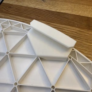 Op de afbeelding: Een wit plastic tafelonderdeel met een geometrisch ontwerp. Het onderdeel heeft een reeks driehoeken en cirkelvormige openingen. Een rechthoekig stuk is aan de bovenkant bevestigd.