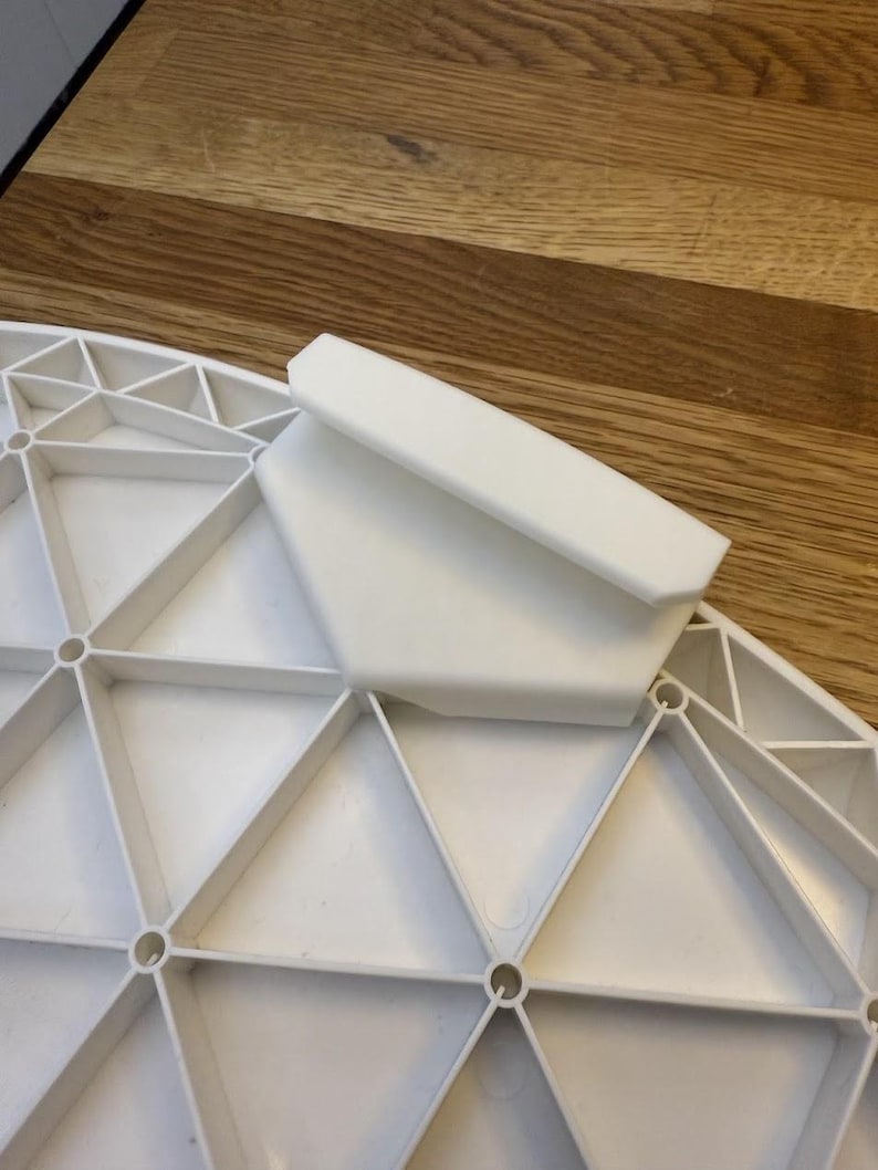 Op de afbeelding: Een wit plastic tafelonderdeel met een geometrisch ontwerp. Het onderdeel heeft een reeks driehoeken en cirkelvormige openingen. Een rechthoekig stuk is aan de bovenkant bevestigd.