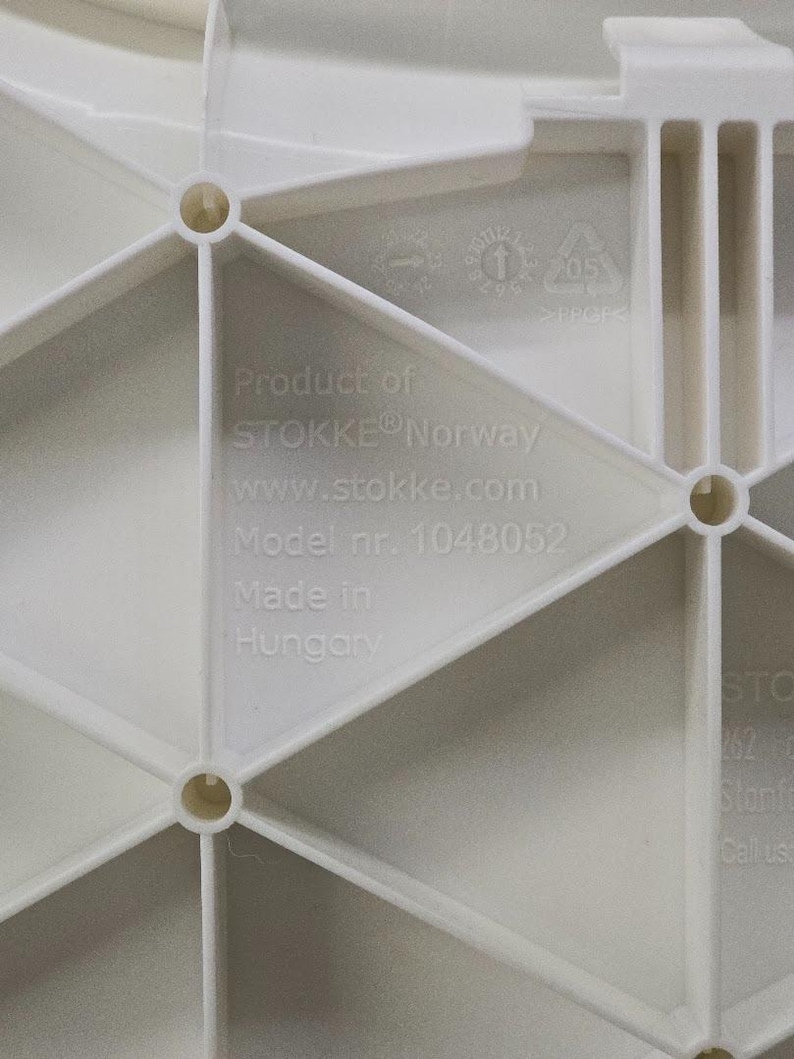 Op de afbeelding: Close-up van een wit kunststof onderdeel met een geometrisch ontwerp. De tekst "Product of STOKKE Norway" en "Model nr. 1048052" zijn zichtbaar. Het onderdeel is gemaakt in Hongarije.