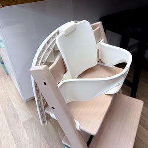 Op de afbeelding: Een witte en natuurlijke houten kinderstoel met een gebogen zitting en rugleuning. De stoel heeft een houten frame en een witte plastic zitting en rug. De stoel is ontworpen voor een kind.