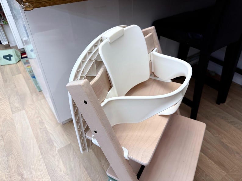 Op de afbeelding: Een witte en natuurlijke houten kinderstoel met een gebogen zitting en rugleuning. De stoel heeft een houten frame en een witte plastic zitting en rug. De stoel is ontworpen voor een kind.