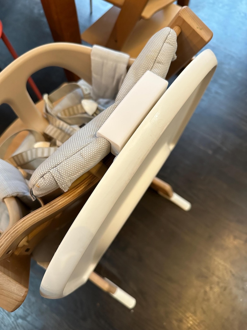 Op de afbeelding: Een lichtgekleurde houten kinderstoel met een wit dienblad en een grijs patroon kussen. De stoel heeft een veiligheidsharnas en een voetensteun. De lade is ongeveer 46 cm breed.